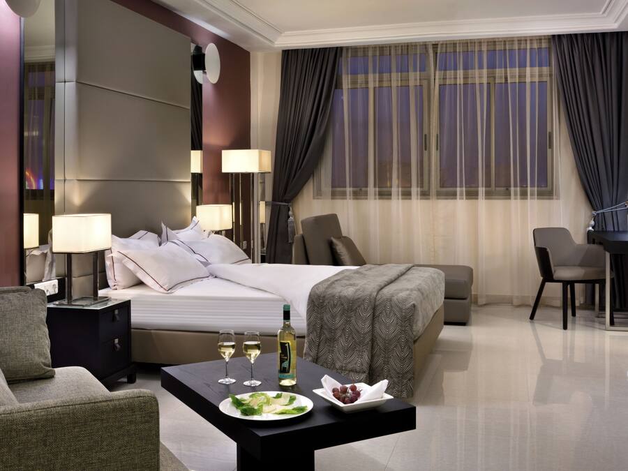 Una habitación de hotel con una cama, mesitas de noche con lámparas, un escritorio, una silla y una mesa de comedor con un plato de comida y copas de vino.