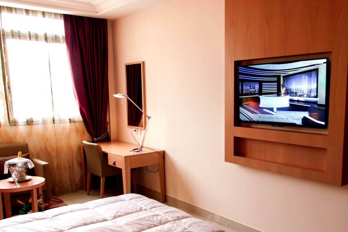 Une chambre d’hôtel comprenant un lit, un bureau avec une chaise, un téléviseur fixé au mur et une fenêtre avec des rideaux.