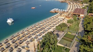 An aerial view of a beach resort with rows of sunbeds and umbrellas, a pool, and a boat docked nearby. - Titanic Luxury Collection Bodrum - All inclusive (Milas)