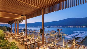 An outdoor dining area with wooden tables and chairs, overlooking a body of water and a distant cityscape. - Titanic Luxury Collection Bodrum - All inclusive (Milas)