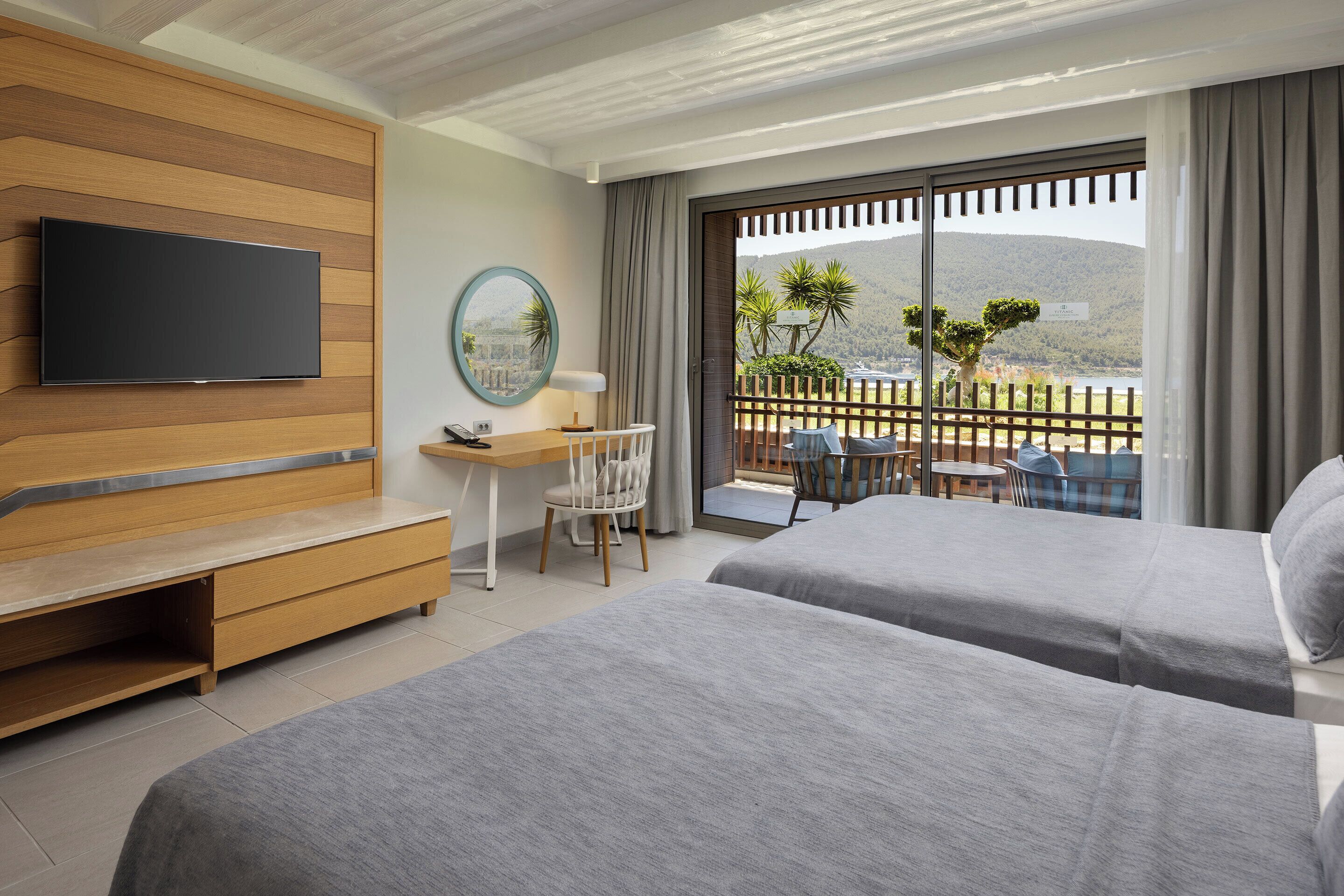 Ein Hotelzimmer mit zwei Betten, einem an einer Holzvertäfelung befestigten Fernseher, einem Schreibtisch mit Stuhl und einem Balkon mit Blick auf eine gebirgige Landschaft.