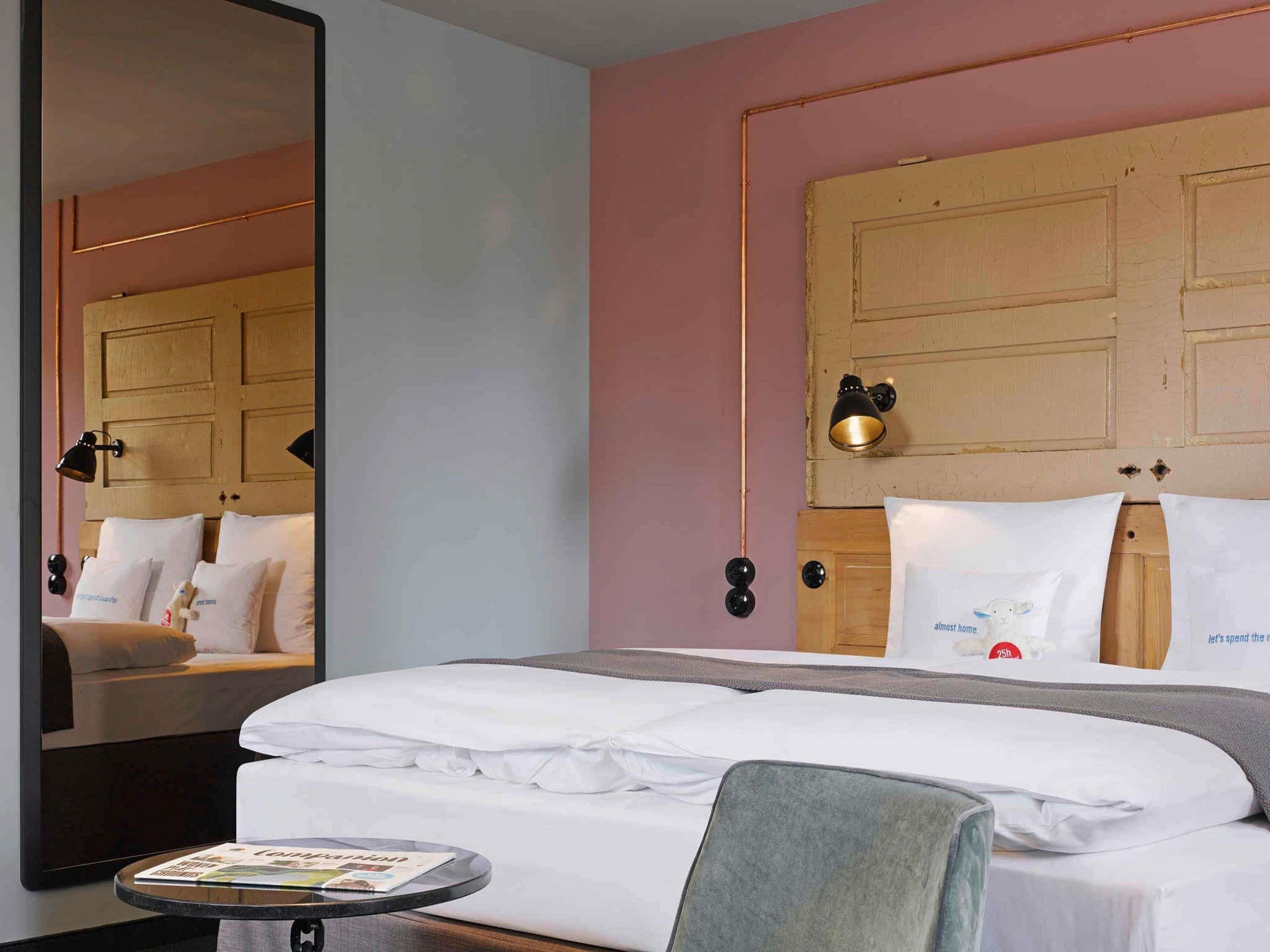 Una camera d'albergo con un letto grande, un comodino con una rivista, uno specchio e una lampada a parete.