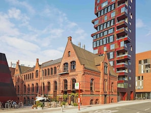 Un bâtiment historique en briques rouges, doté d’arcs en plein cintre au niveau des fenêtres et d’un toit plat, situé à côté d’un immeuble moderne de grande hauteur.