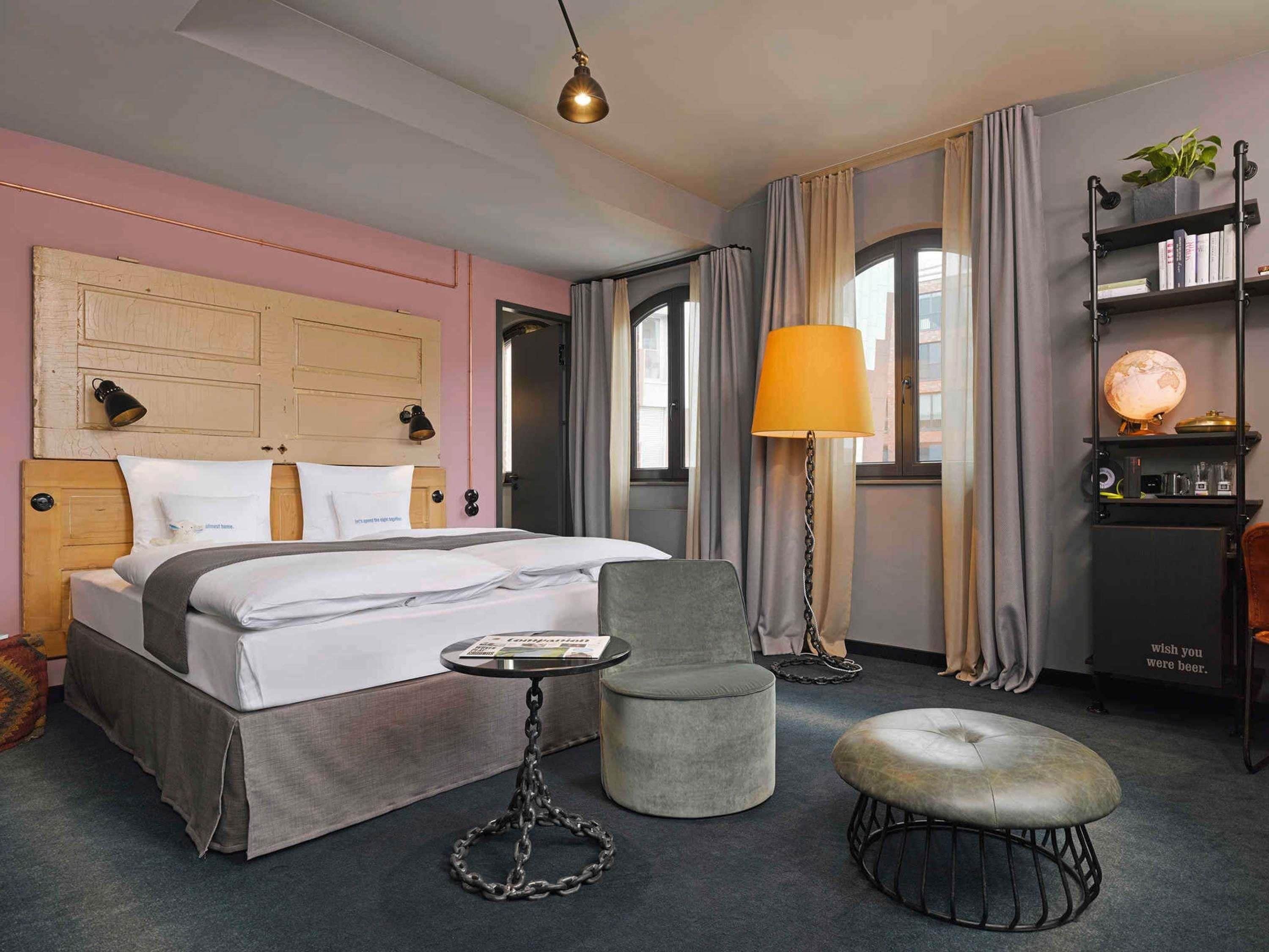 A hotel room with a bed, bedside table, chair, ottoman, lamp, and a shelf with books and a globe.
