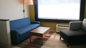 Une pièce avec un canapé bleu, une table basse en bois, un fauteuil gris, une grande fenêtre avec des stores, un lampadaire et un radiateur.