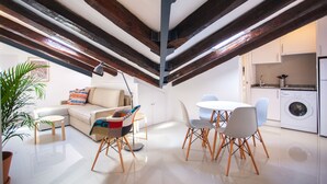 A modern loft apartment with exposed wooden beams, a dining area with a round table and chairs, a sofa with colorful cushions, a small kitchen with a washing machine, and a potted plant. - Apartment in Malasaña   (Madrid)