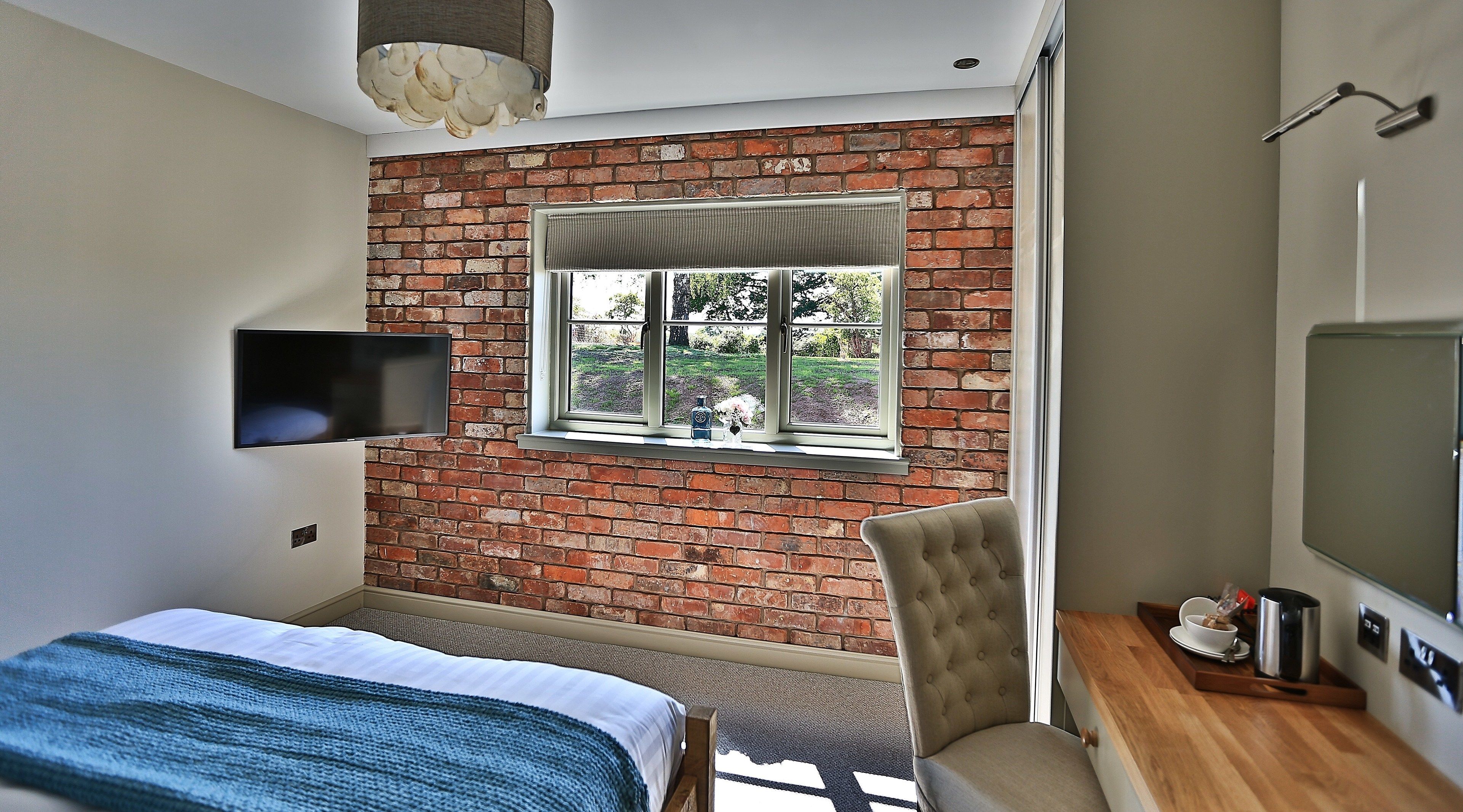 A bedroom with a brick wall, a window, a bed, a chair, a TV, and a wooden table with a kettle.