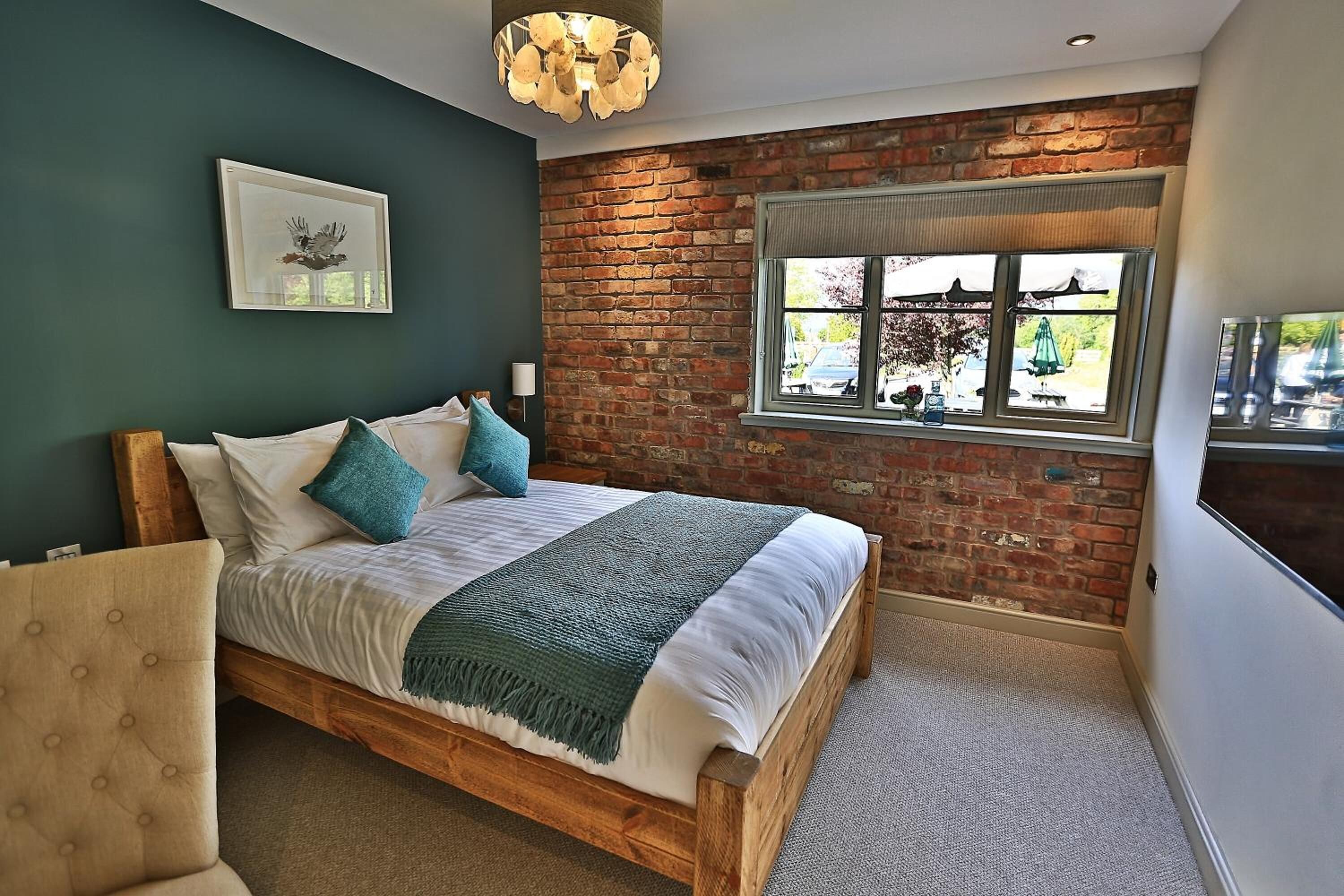 A bedroom with a wooden bed, a brick wall, a window with a view, and a ceiling light fixture.