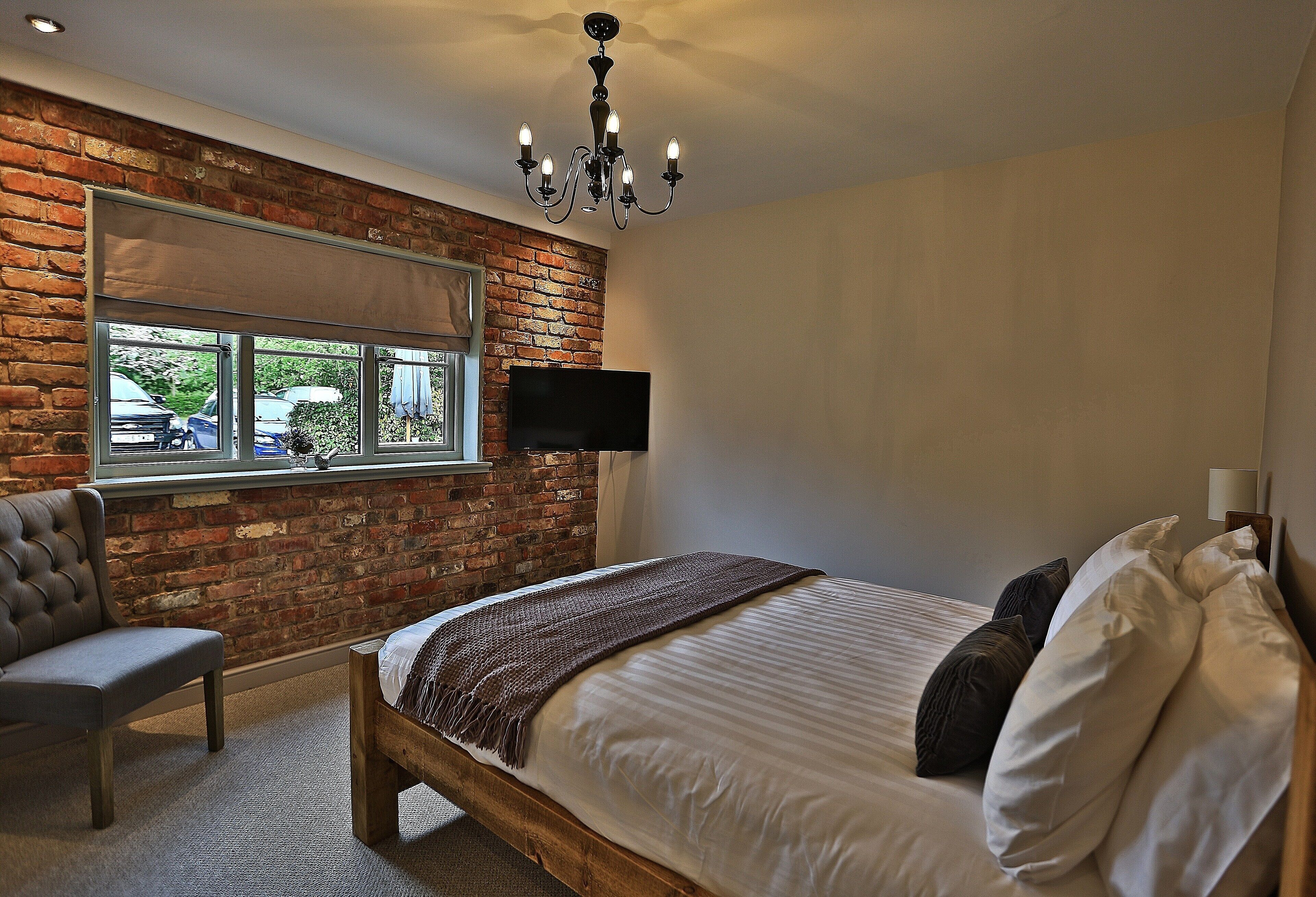 A bedroom with a brick wall, a large window, a flat-screen TV, a bed with white linens, and a chandelier.