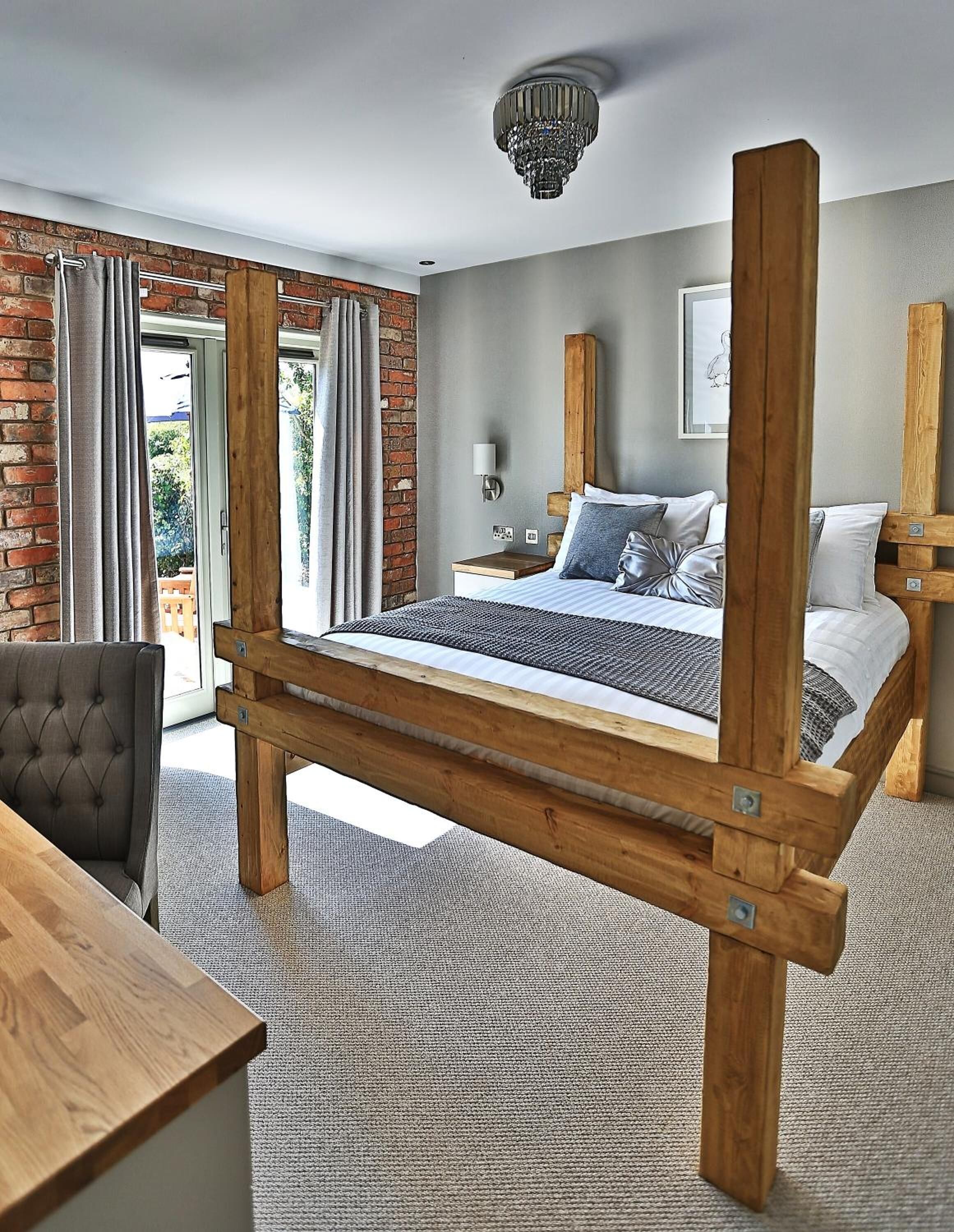 A bedroom with a wooden four-poster bed, a grey carpet, a grey upholstered chair, and a wooden table.