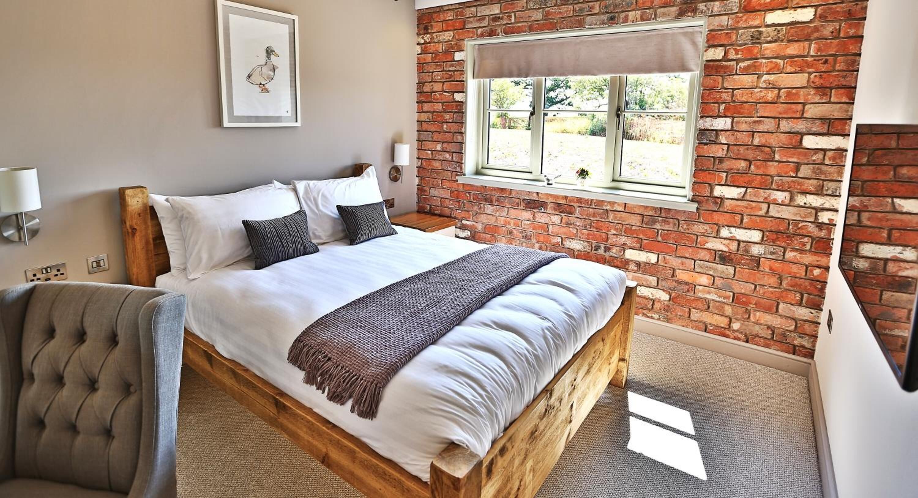 A bedroom with a wooden bed, a brick wall, a window with a curtain, and a wall-mounted lamp.