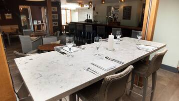 Un restaurante con una mesa de mármol preparada para cuatro personas, sillas de mimbre y una zona de bar al fondo.