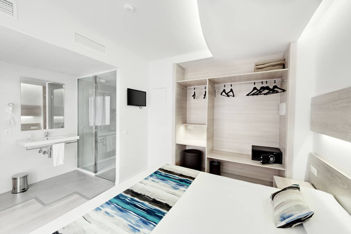 Une chambre d’hôtel moderne dotée d’une douche en verre, d’une coiffeuse blanche, d’un lit avec un tapis à rayures et d’un dressing où sont suspendus les vêtements.