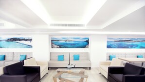 Un hall d’hôtel moderne avec un espace salon central, une table basse en verre et de grandes peintures représentant des paysages côtiers accrochées aux murs.