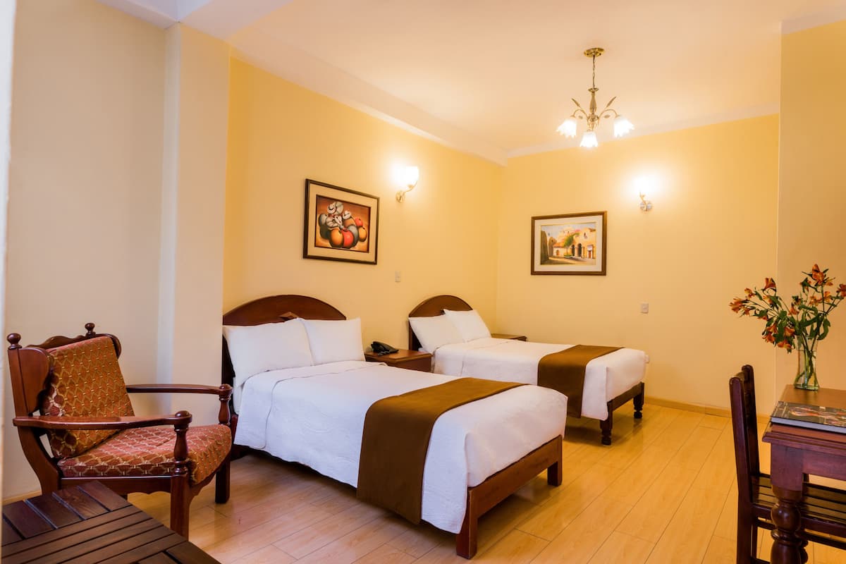 Habitación de hotel con dos camas, una silla de madera, una mesa y cuadros colgados en la pared.