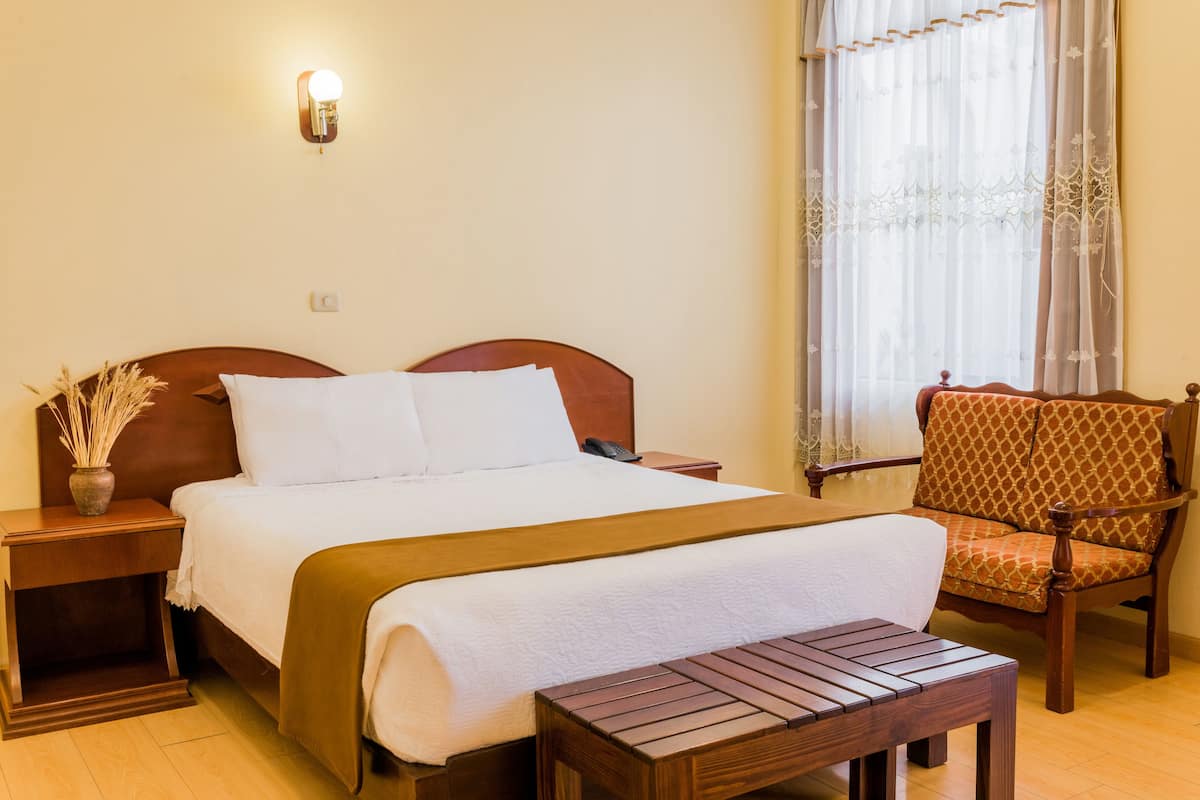 Una habitación de hotel ordenada con una cama grande, una mesita de noche de madera, una mesilla auxiliar, una silla y una ventana con cortinas traslúcidas.