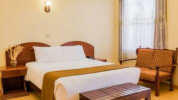 A neatly arranged hotel room with a large bed, a wooden nightstand, a bedside table, a chair, and a window with sheer curtains.