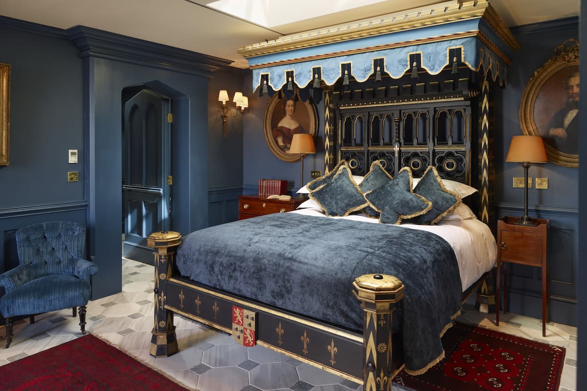 A bedroom with a four-poster bed, a red and black patterned rug, a blue armchair, and a wooden nightstand with a lamp.