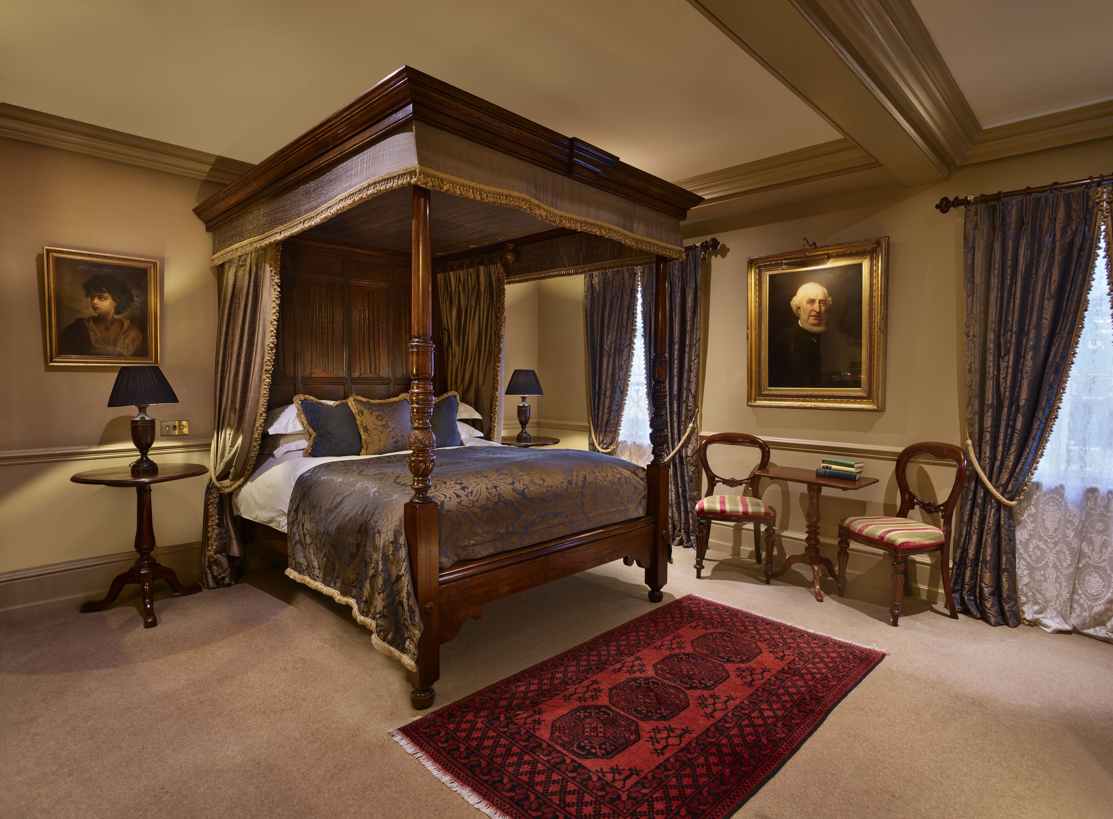 A four-poster canopy bed with a red patterned rug, two bedside tables with lamps, and a chair with a striped cushion.