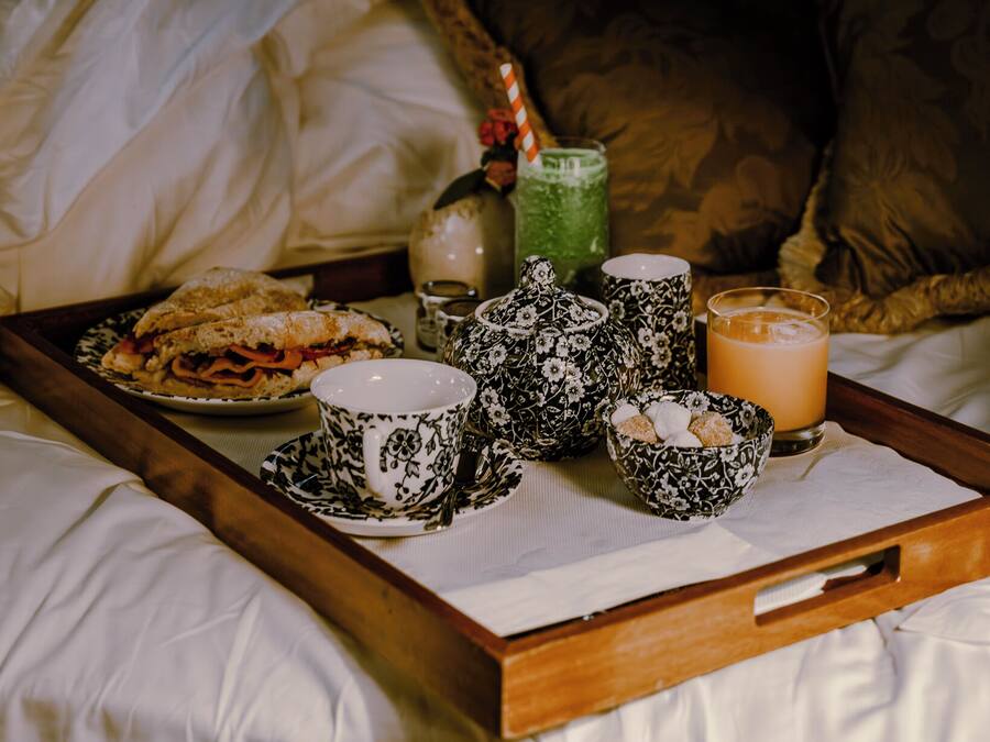 A bed with a tray of breakfast items including a sandwich, tea cups, a teapot, and a glass of orange juice.