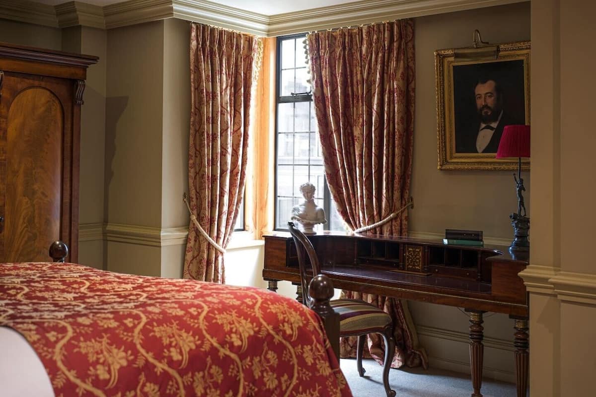 A room with a large bed, a desk with a bust sculpture, a chair, and a framed portrait on the wall.