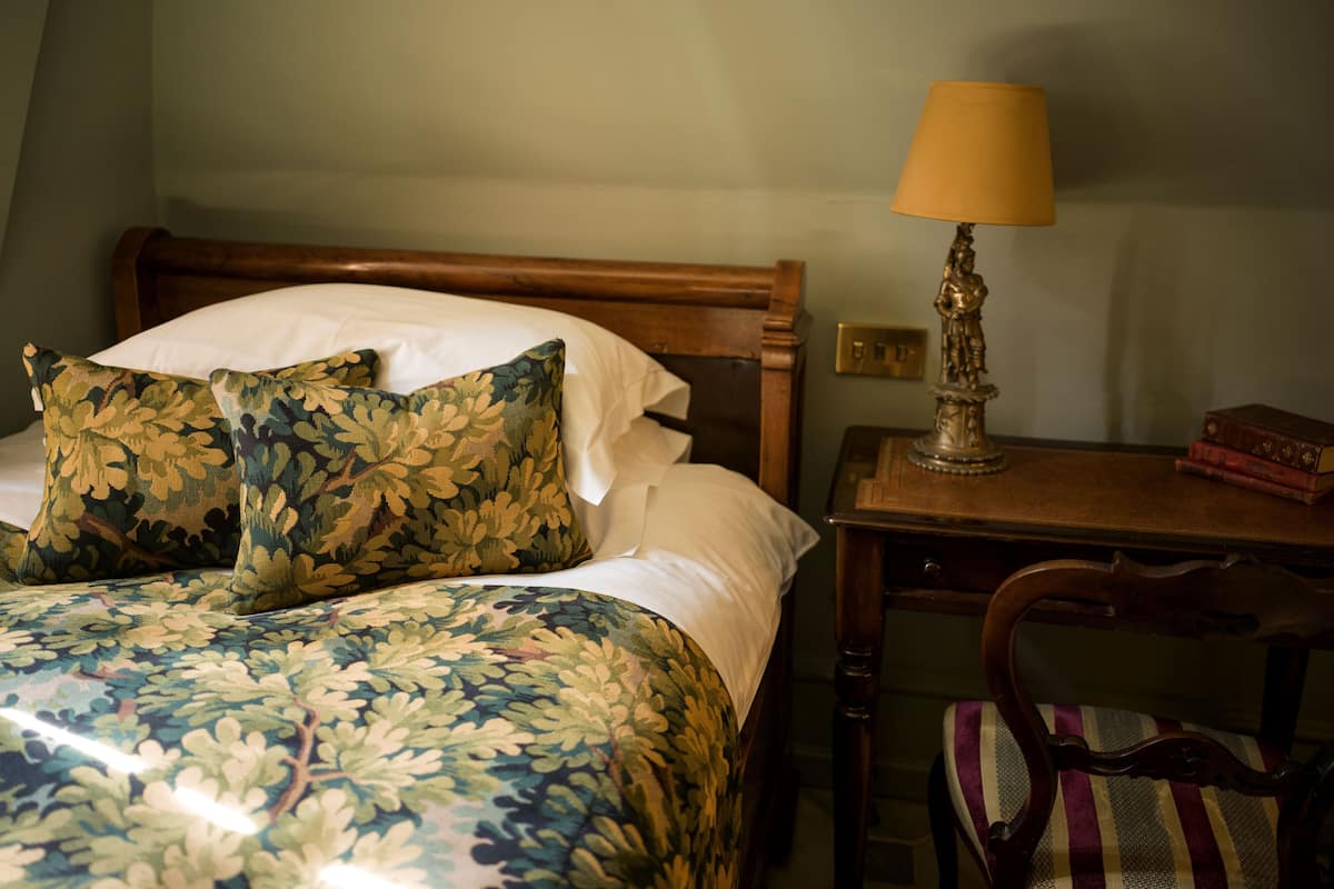 A bed with floral bedding, a wooden headboard, a bedside table with a lamp, and a chair with a striped cushion.