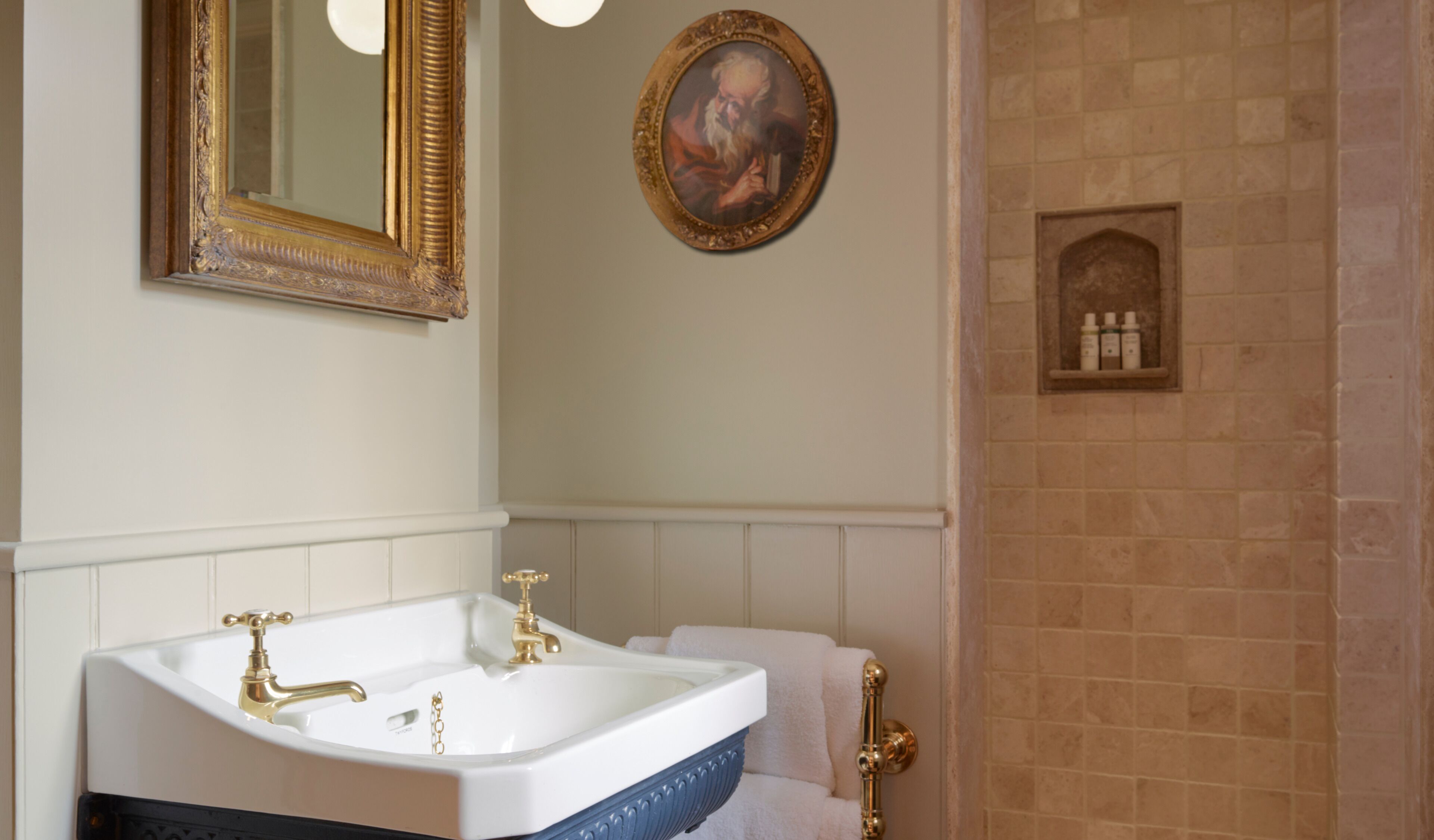 Une salle de bain avec un lavabo blanc, des robinets dorés, un miroir avec un cadre doré et un tableau accroché au mur.