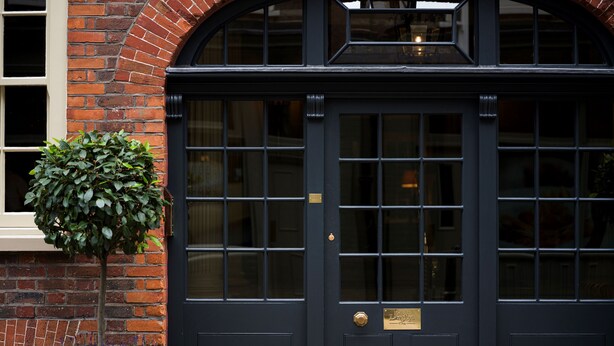 A black double door with a brass plaque, a small tree, and a brick wall. - Batty Langley's (London)