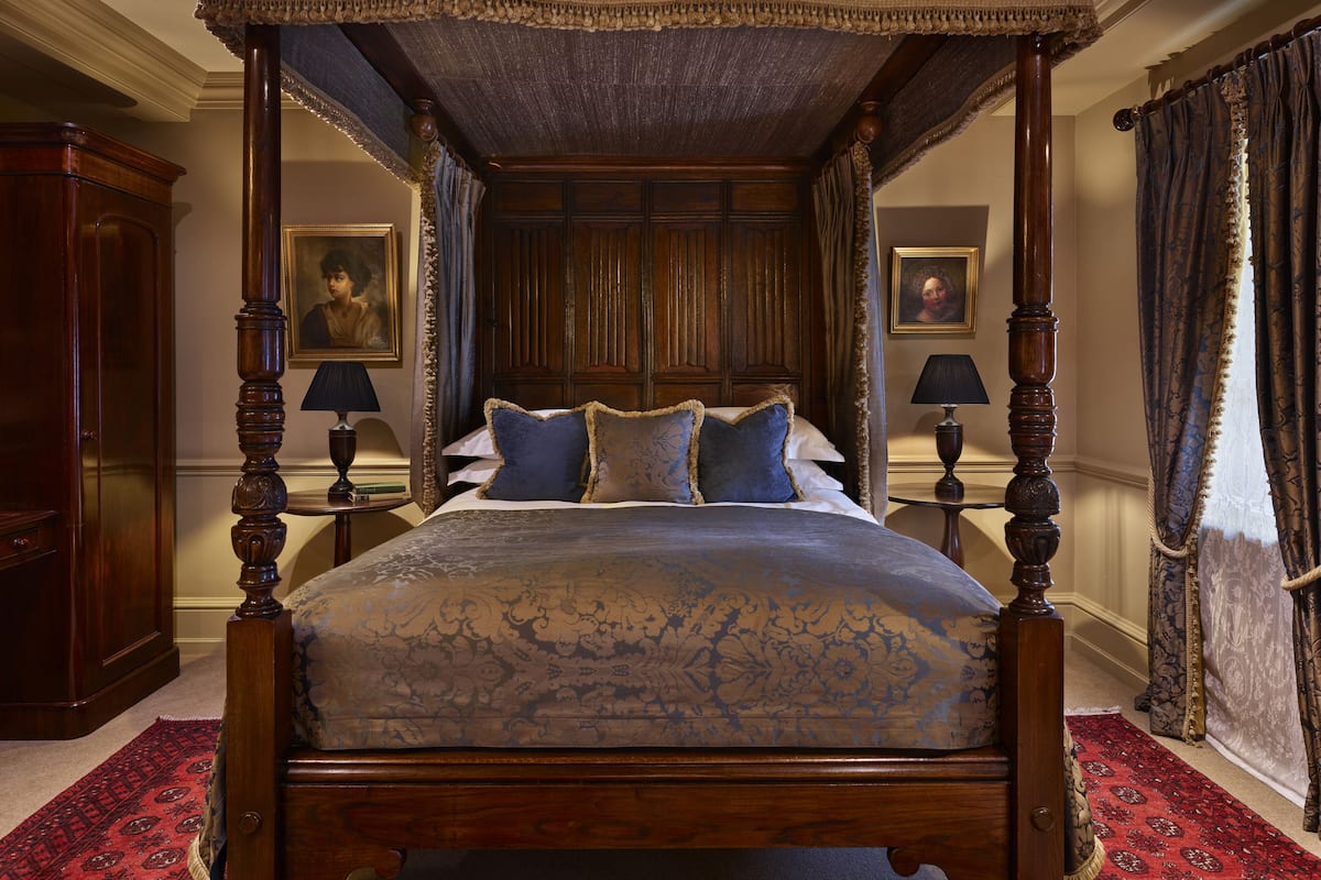 A four-poster canopy bed with a floral bedspread, two nightstands with lamps, and a red patterned rug.