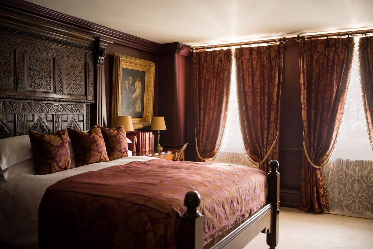A classic four-poster bed with a patterned bedspread, a nightstand with a lamp, and a framed painting on the wall.