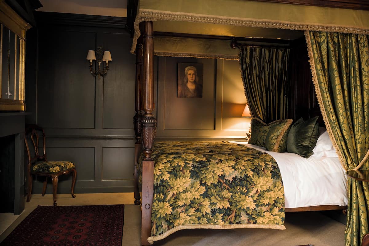 A four-poster bed with a floral bedspread, a green canopy, and a nightstand with a lamp. There is a chair with a floral cushion and a red rug on the floor.