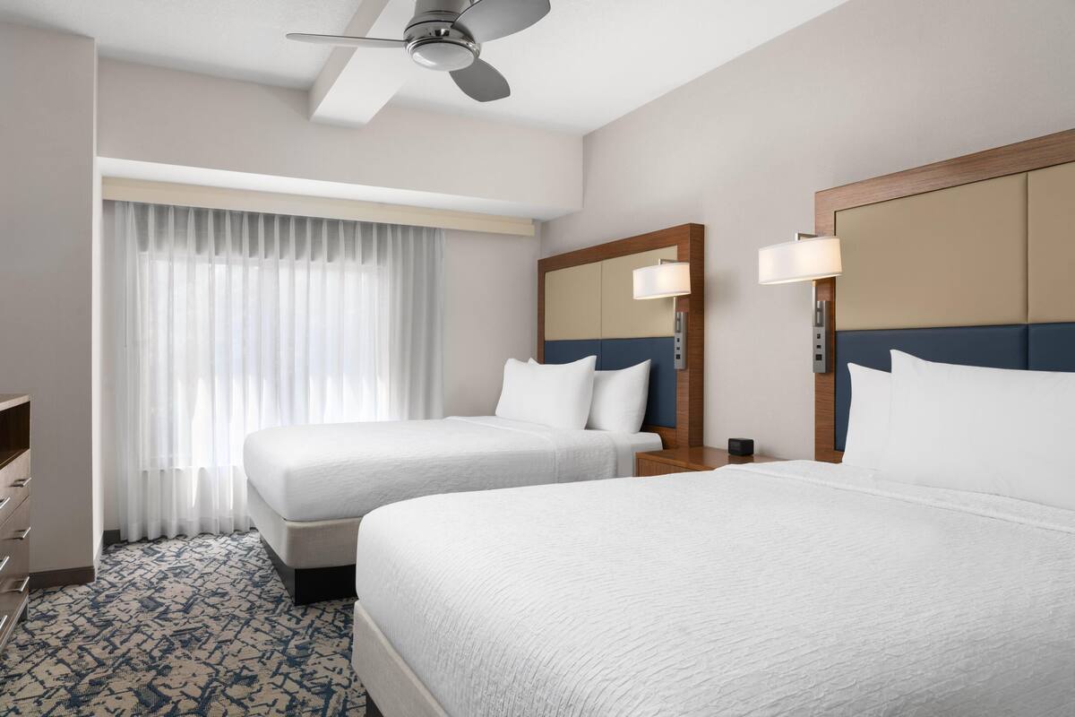 Quarto de hotel com duas camas, um ventilador de teto, uma janela grande com cortinas, uma mesa de cabeceira com abajur e um armário.