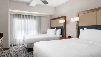Une chambre d’hôtel avec deux lits, un ventilateur de plafond, une grande fenêtre avec des rideaux, une table de chevet avec une lampe et une commode.