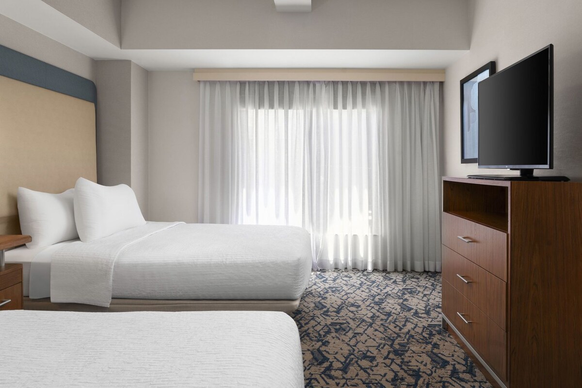 A hotel room with two beds, a television on a wooden cabinet, and a large window with sheer curtains.