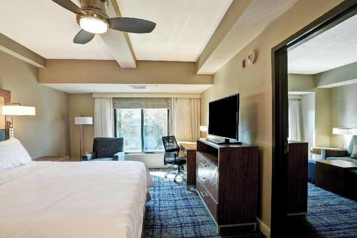 Una habitación de hotel con cama, escritorio, silla, televisión y un ventanal grande.