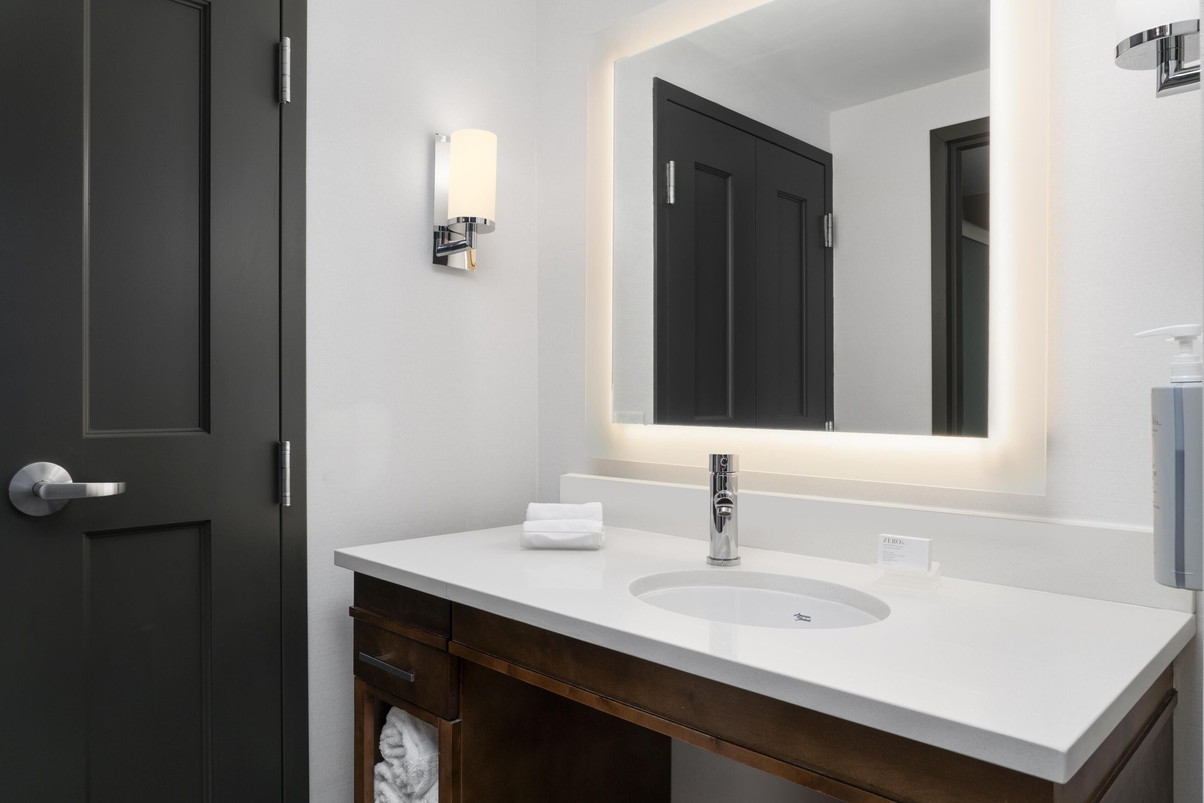 A modern bathroom with a white sink, dark wooden vanity, and a large mirror with integrated lighting.