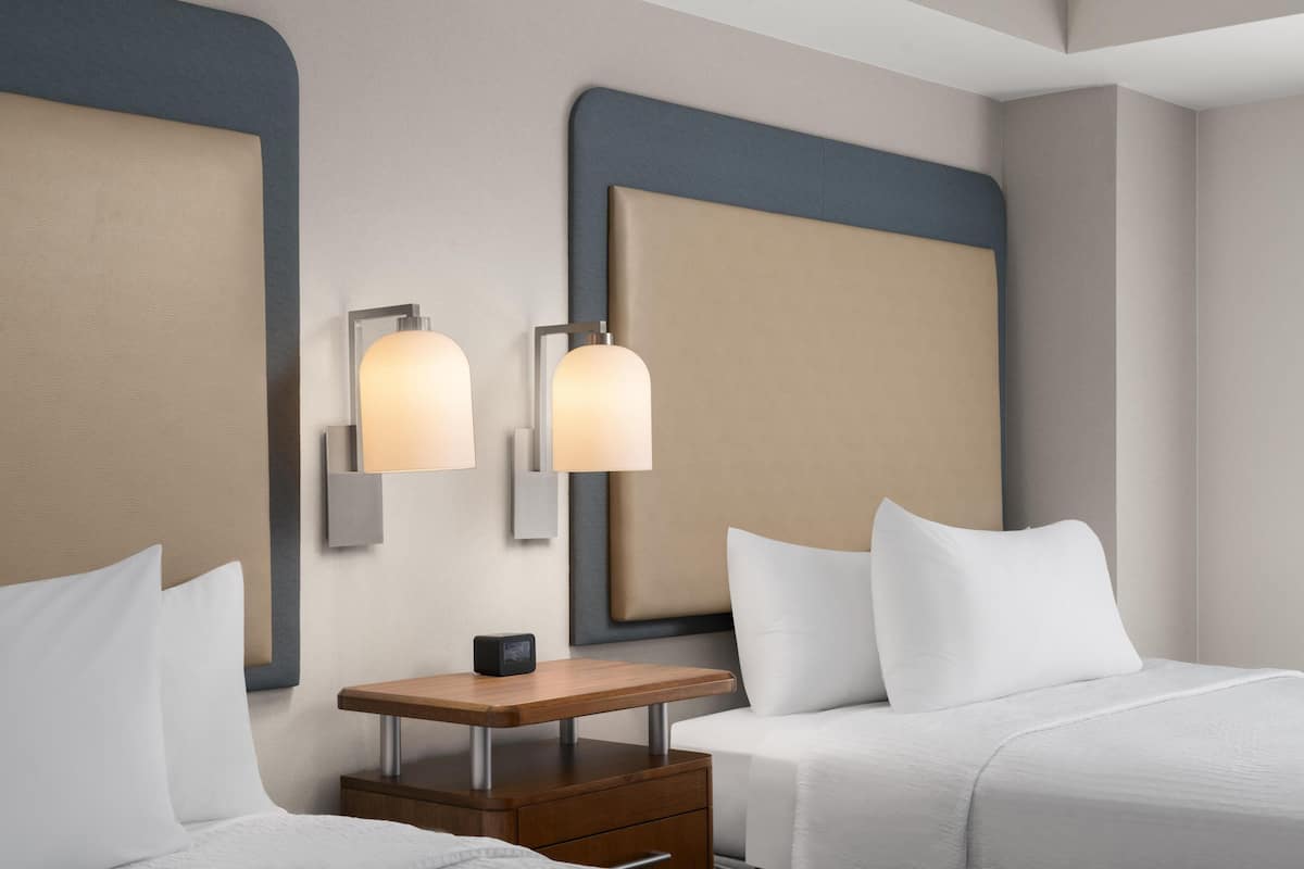 Quarto de hotel com duas camas, uma mesinha de cabeceira e luminárias fixadas na parede.