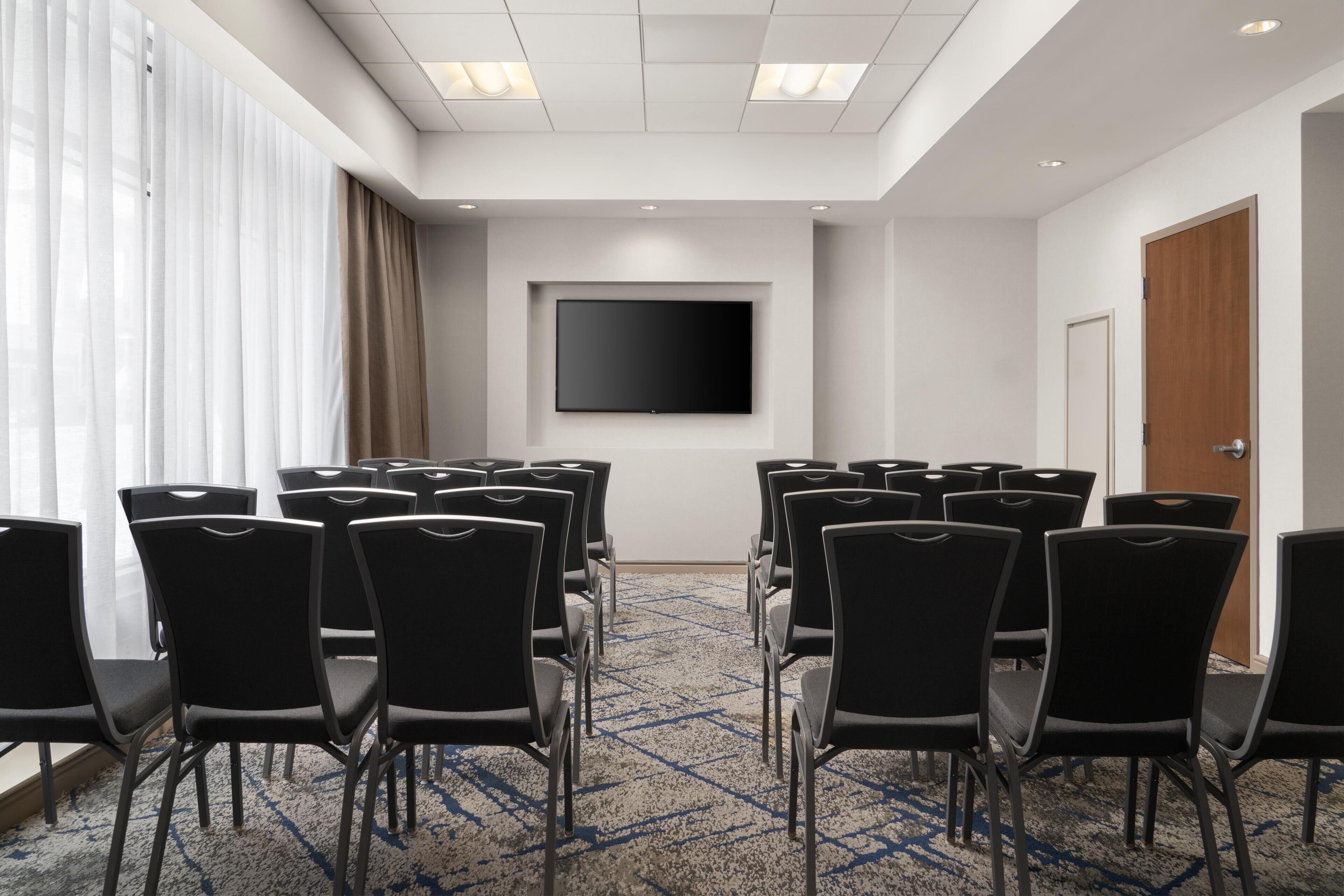 A conference room with a flat-screen TV, rows of black chairs, a carpeted floor, and a large window with curtains.