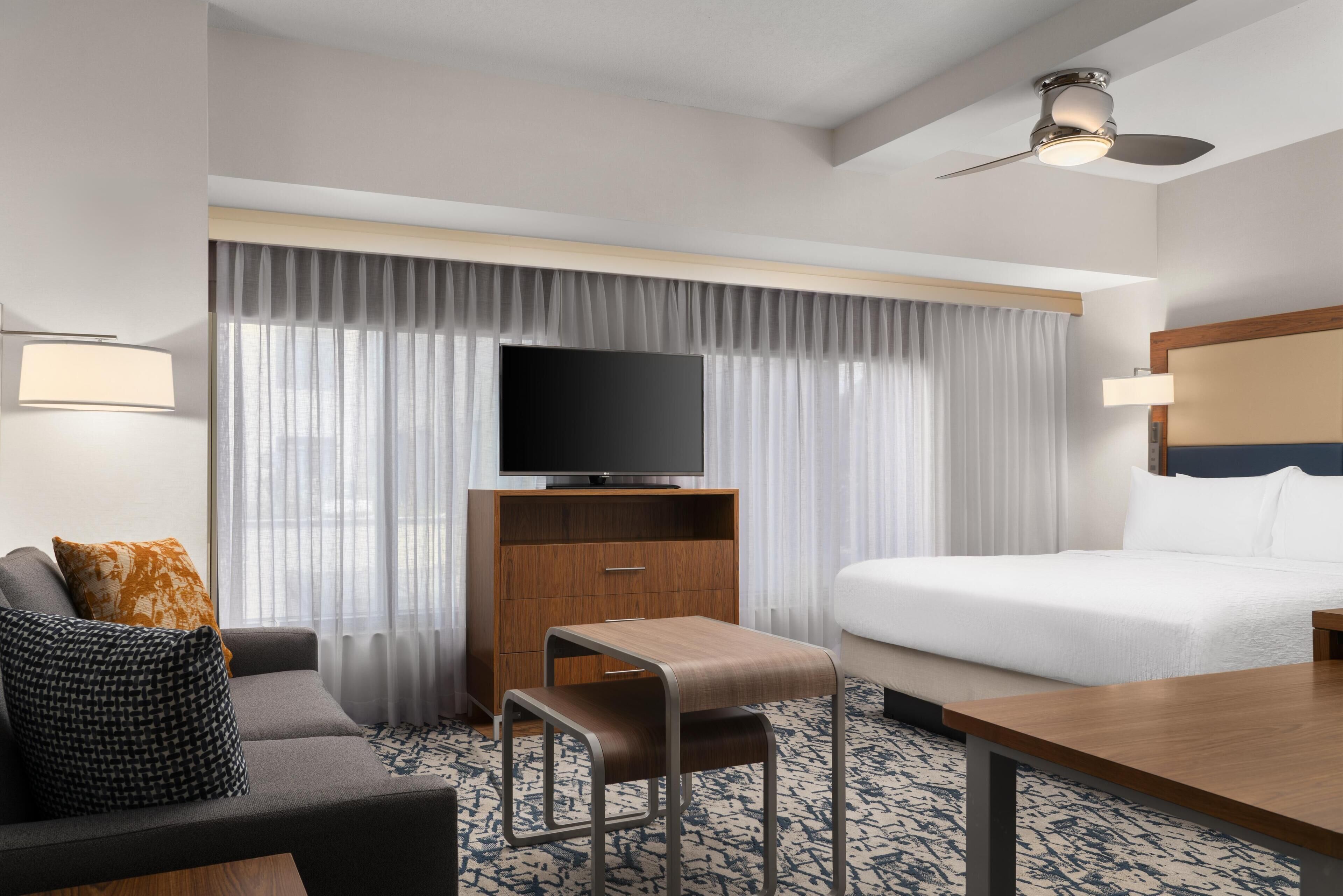 Ein Hotelzimmer mit einer Couch, einem kleinen Tisch, einem Fernseher auf einer Holzkommode, einem Bett und einem Deckenventilator.