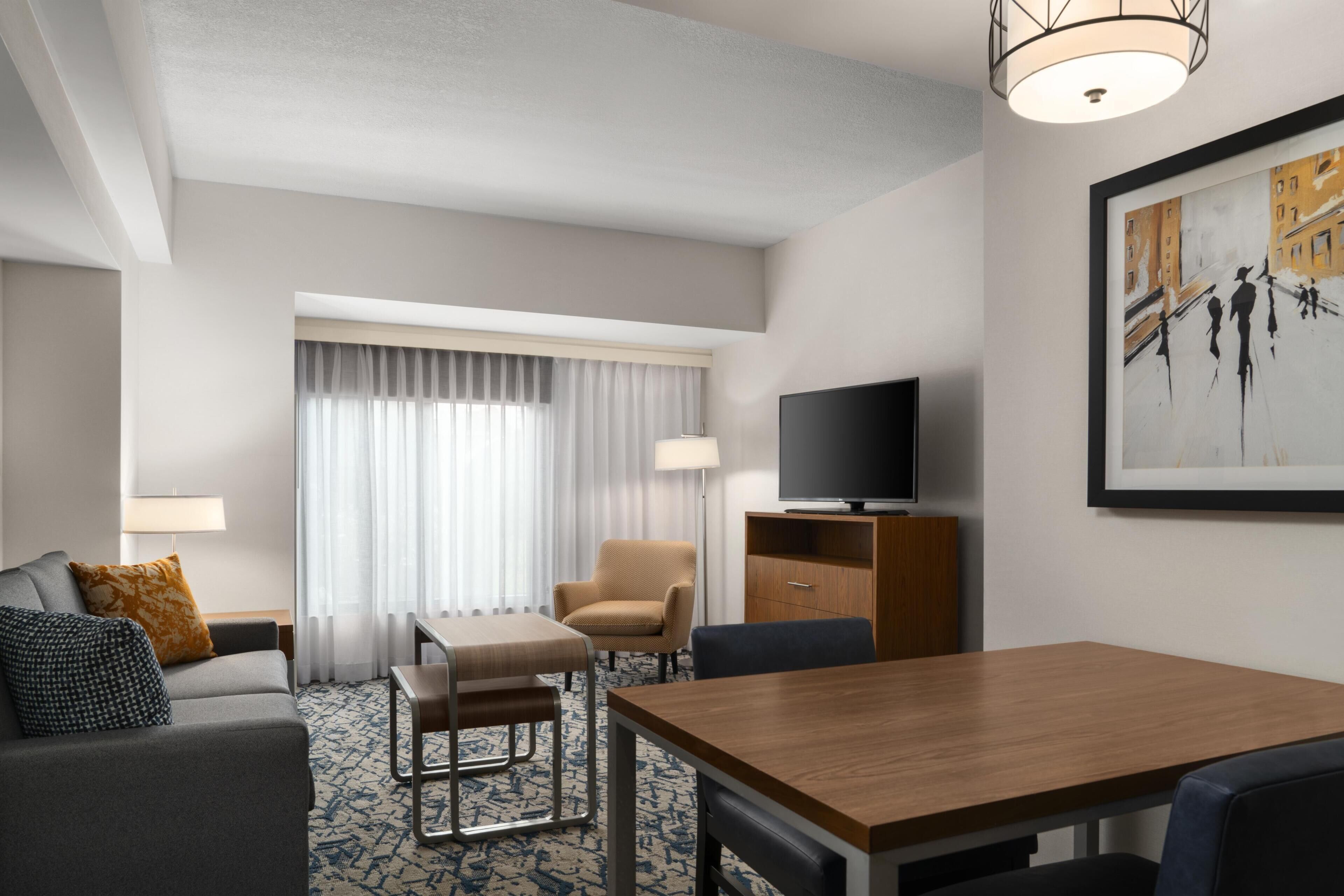 A hotel room with a sofa, armchair, dining table, TV, and artwork on the wall.