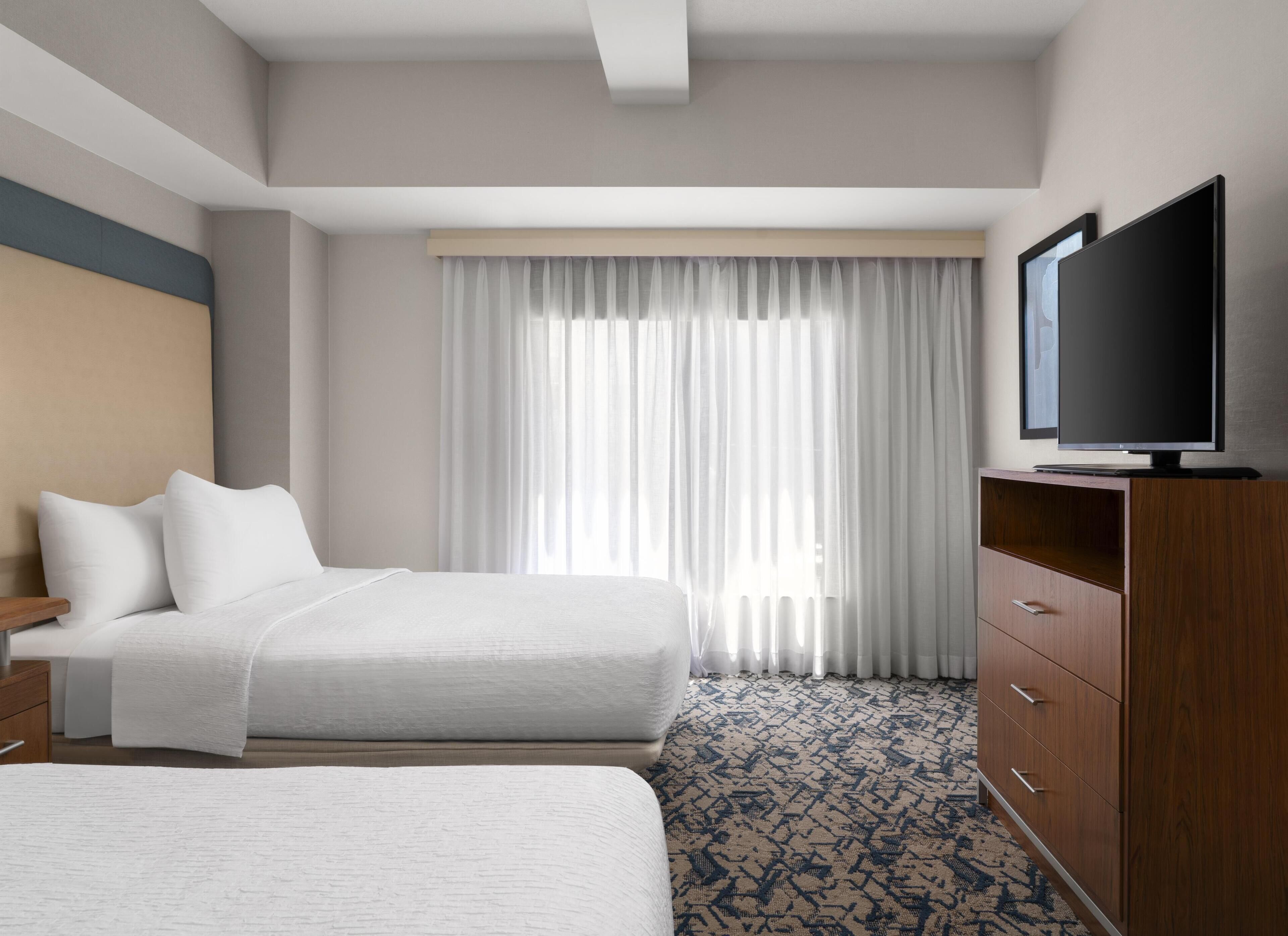 A hotel room with a bed, a TV on a wooden cabinet, and a large window with sheer curtains.