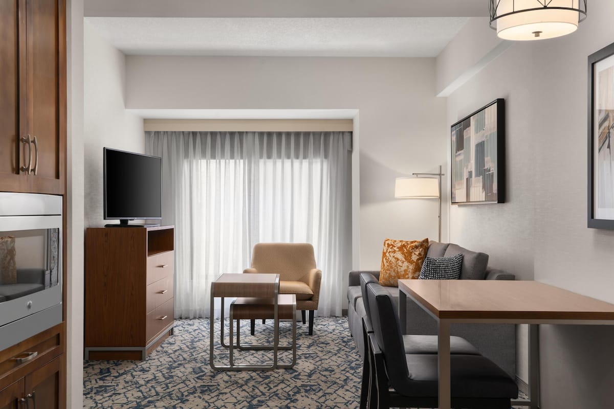 Quarto de hotel com televisão, sofá, mesa pequena, cadeira, escrivaninha, abajur e um quadro na parede.
