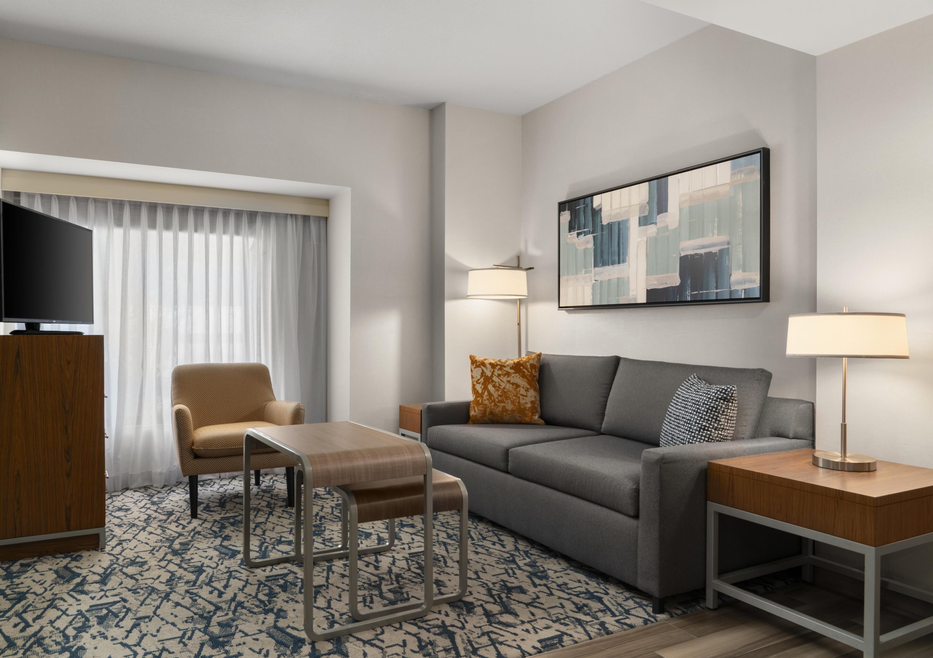 Una habitación de hotel con sofá, sillón, televisor, dos lámparas y un cuadro en la pared.