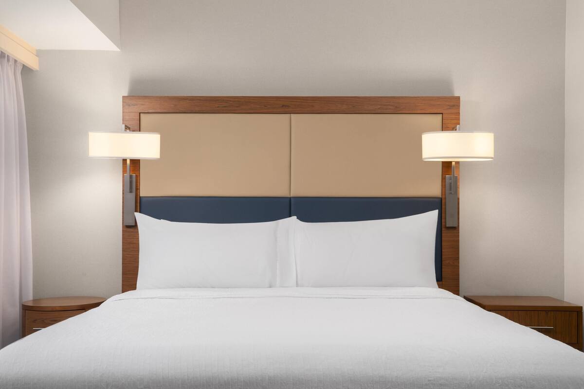 Quarto de hotel com uma cama grande, duas luminárias de cabeceira, painel de cabeceira de madeira e um criado-mudo em cada lado.
