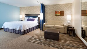 A hotel room with a bed, a sofa, an ottoman, a small table, and a lamp. - Home2 Suites by Hilton Milwaukee Airport (Milwaukee)