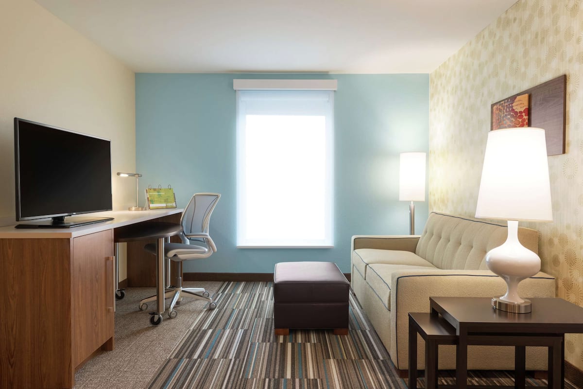 Een hotelkamer met een flatscreen televisie, een bureau met stoel, een bank, een lamp en een raam met jaloezieën.