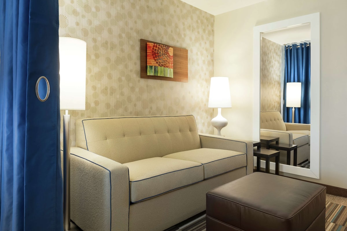 Hotelkamer met een beige bank, een klein tafeltje, een spiegel en een blauw gordijn.