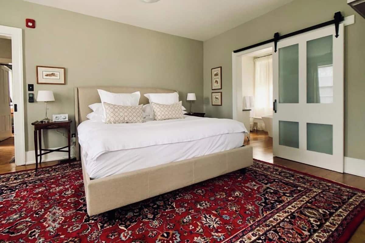 A spacious bedroom with a large bed, a red patterned rug, and a sliding glass door to the right.