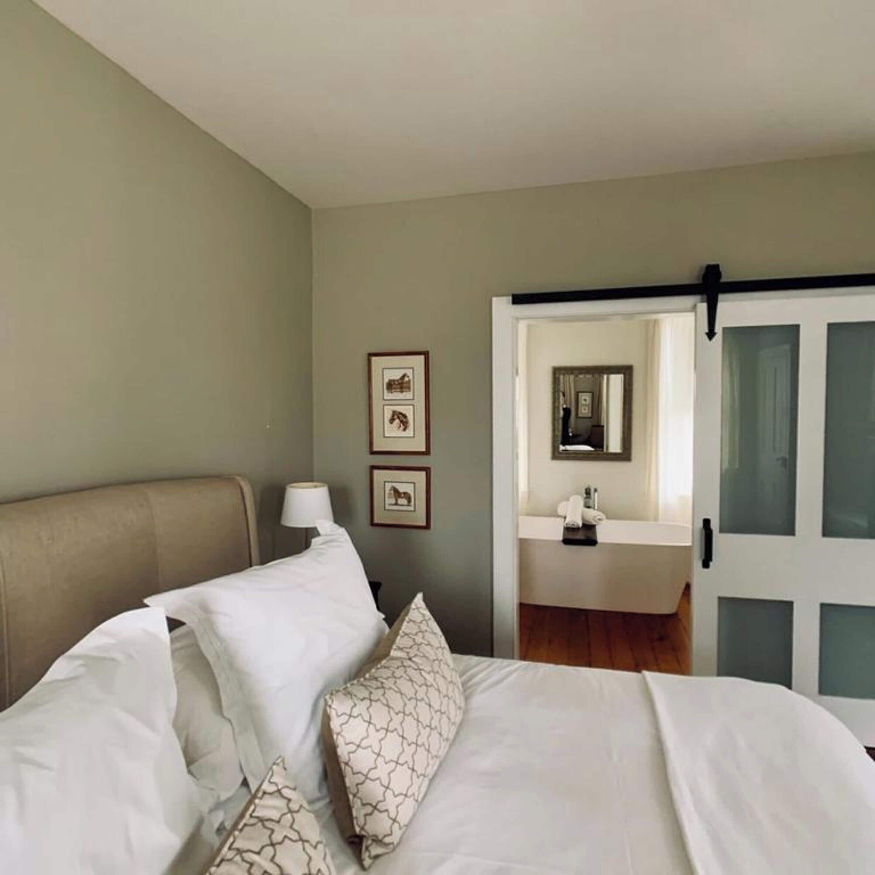 A bedroom with a bed, a nightstand, a lamp, and a bathroom visible through an open door.