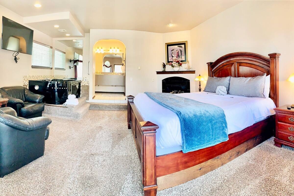 A bedroom with a wooden bed, a sitting area with a sofa and armchair, a fireplace, and a hallway leading to a bathroom.